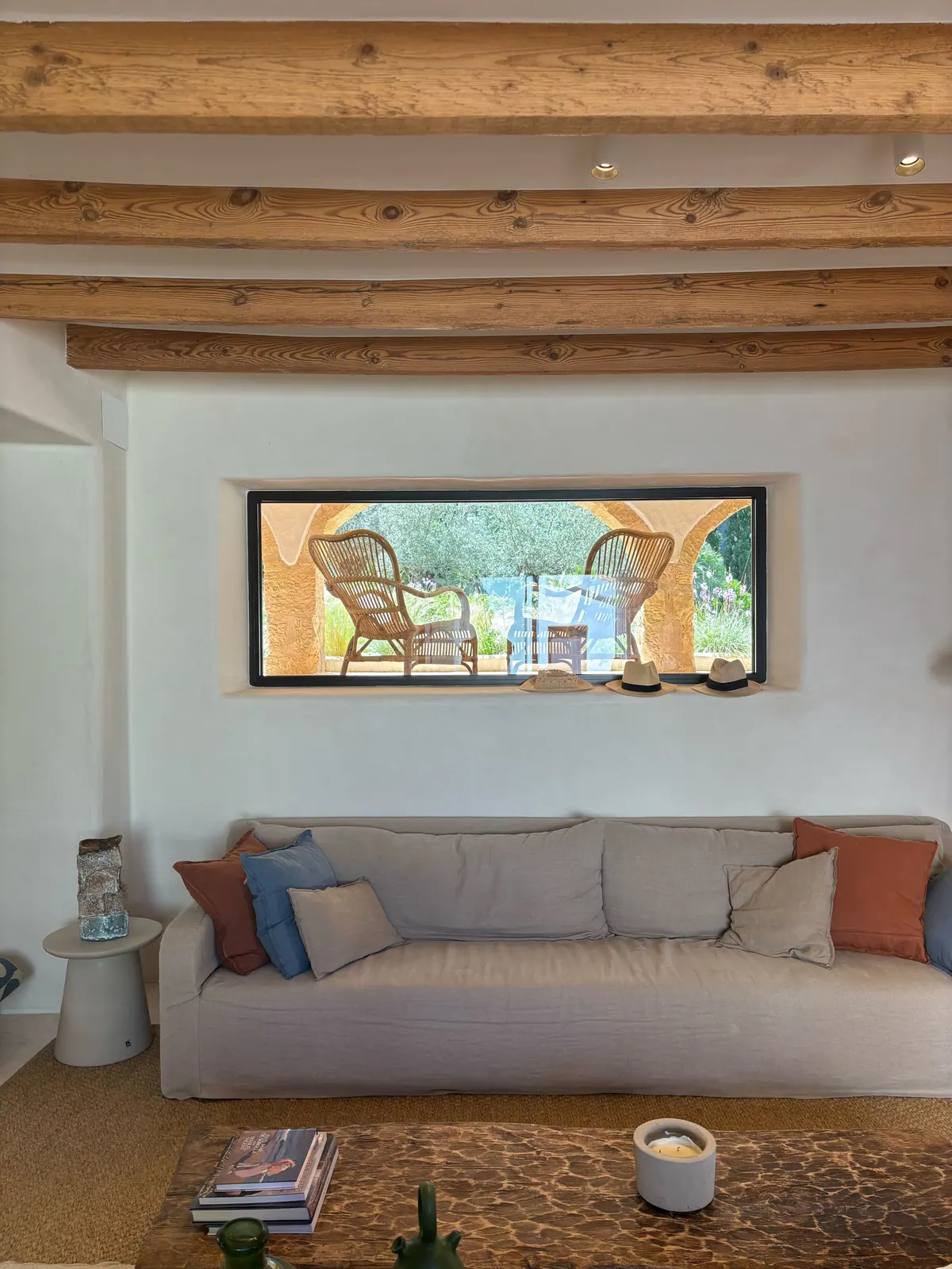 A living room with timber beams, a linen sofa and an interior window framing a terrace