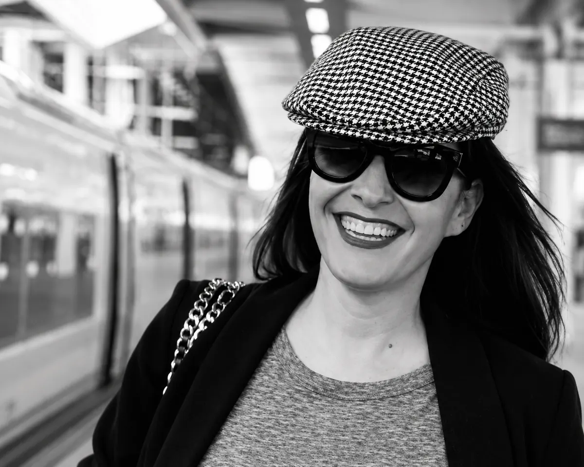 Jessica Bataille smiling on a station platform, in a houndstooth cap and dark glasses