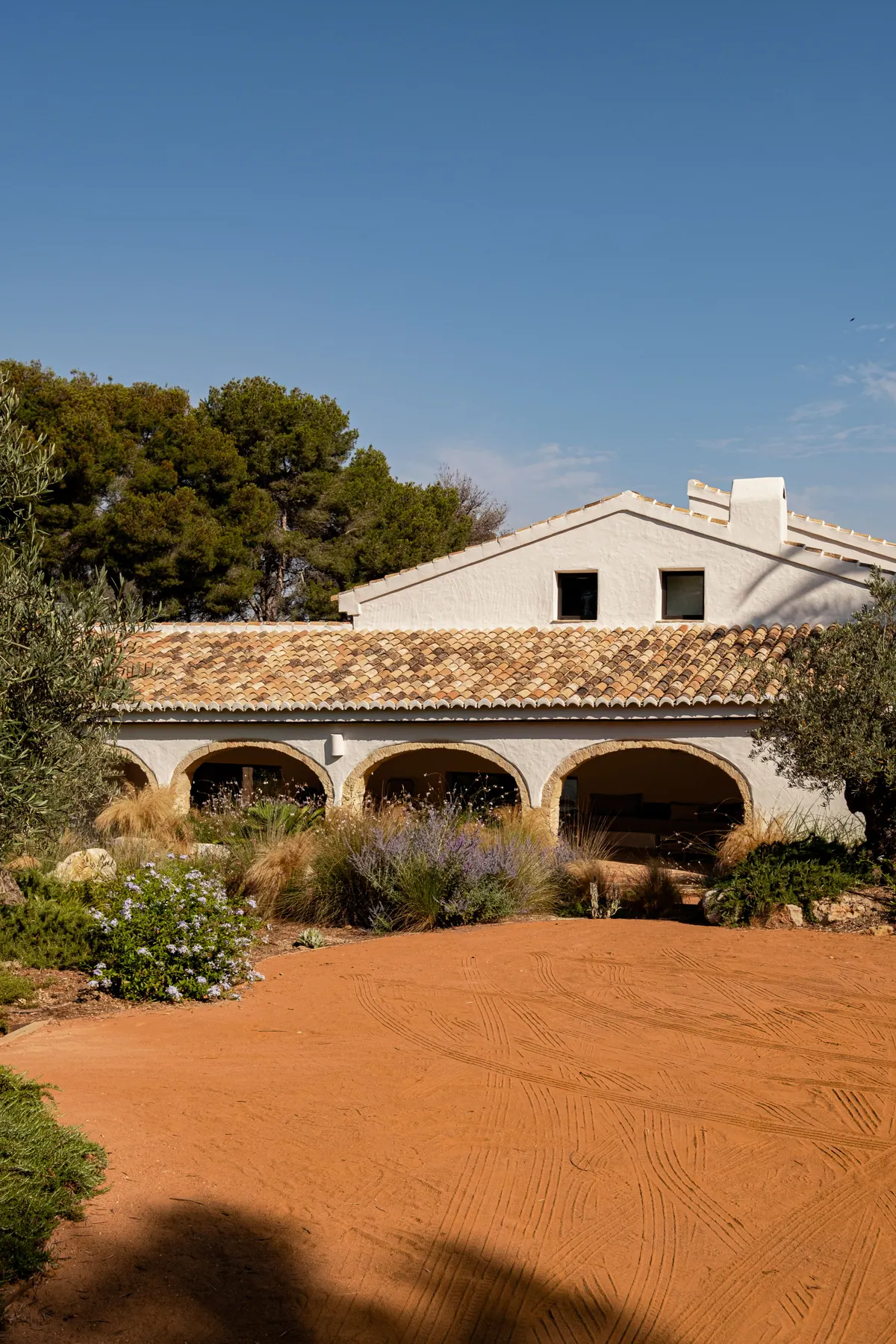 A whitewashed masía with a row of arches and a tiled roof behind a dry garden