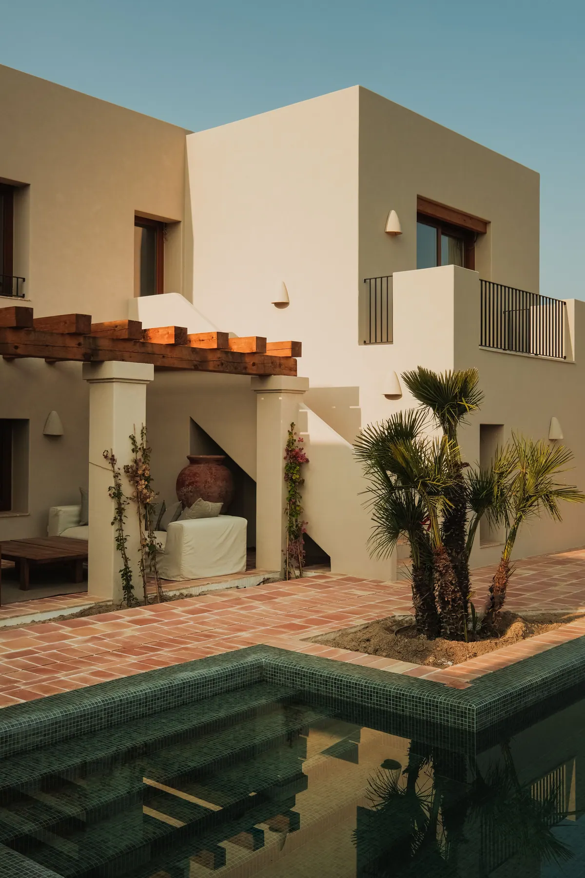 A courtyard with terracotta paving, a green-mosaic pool, palms and a timber pergola