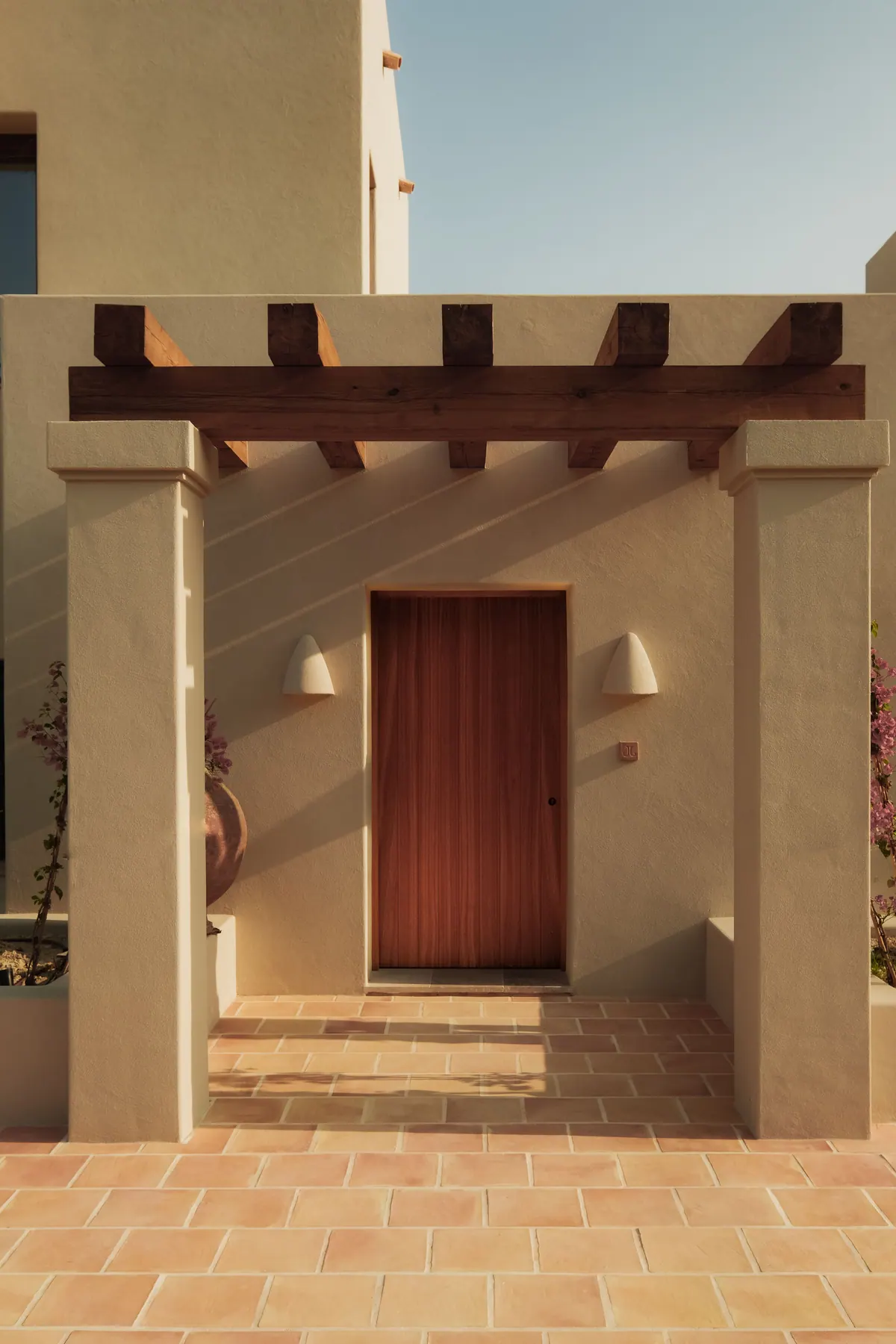 A timber door under a beamed pergola, framed by rendered pillars and bougainvillea